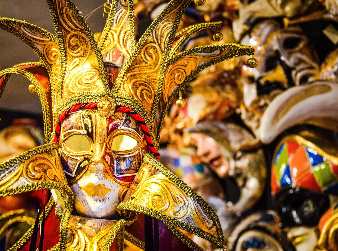 A gilded jester mask with spiralling gold points and a white face, set against a crowd of other Venetian masks