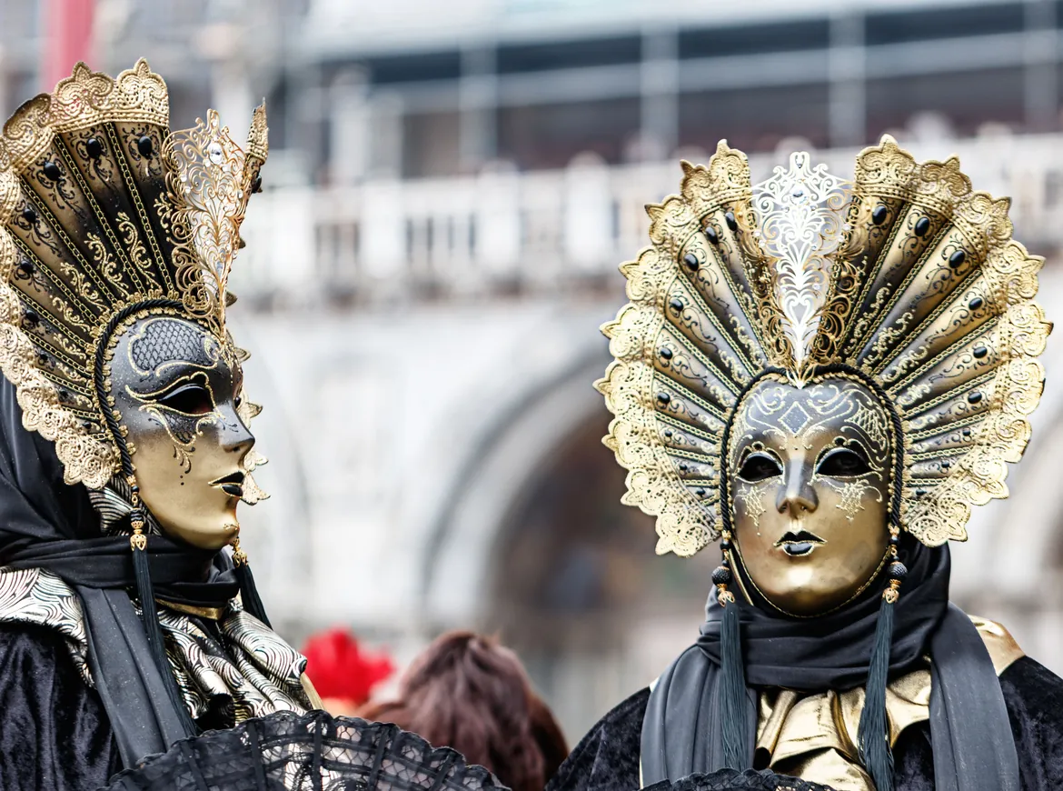 Two people in dark costumes and tall fan-shaped gold headdresses with gilded masks, in front of a pale arcaded building